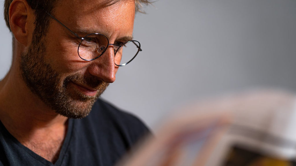 A man wearing Danish designed glasses is engrossed in reading a newspaper.